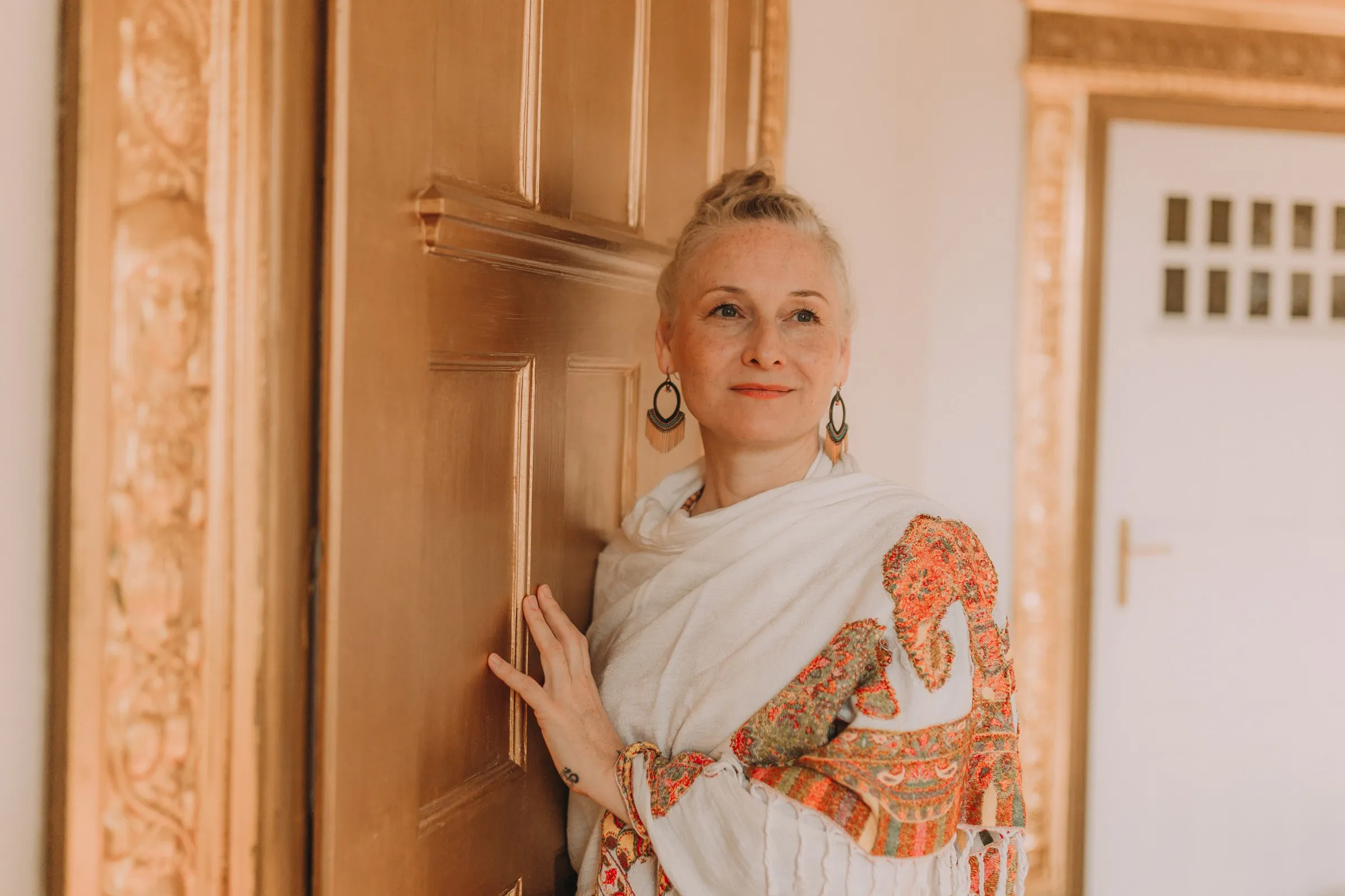 Kathrin Seemann lehnt in einem weißen Tuch an einem goldenen Türrahmen der YogaLounge